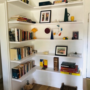 May include: White corner shelves with books, a lamp, a mobile, a vase, a picture frame, and a small wooden stool. The shelves are filled with books and decorative items. The stool is made of wood and has a woven top.