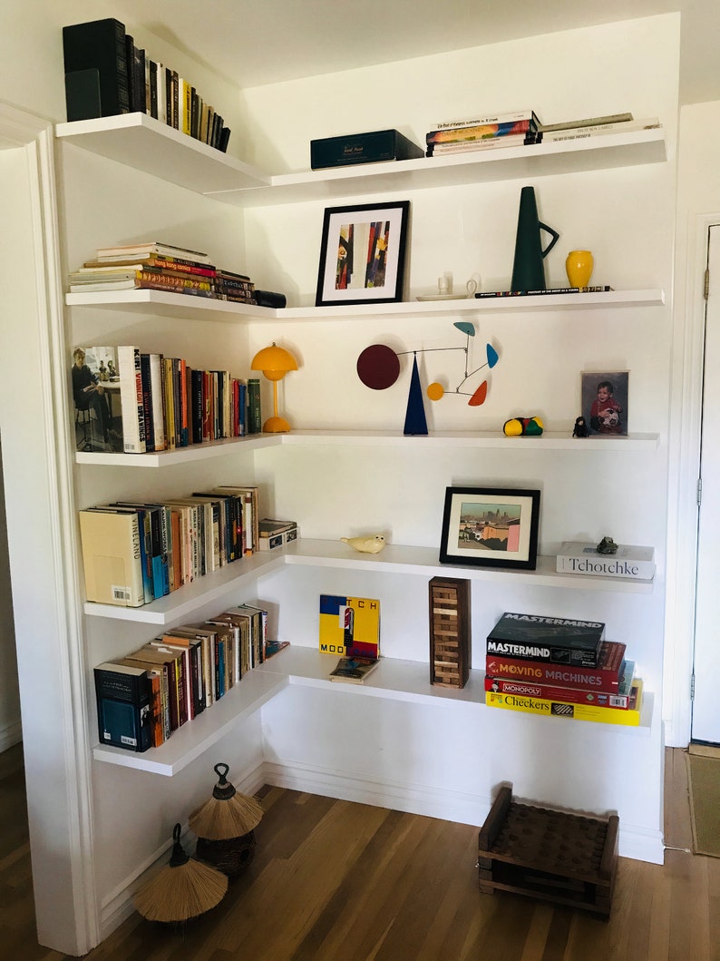 May include: White corner shelves with books, a lamp, a mobile, a vase, a picture frame, and a small wooden stool. The shelves are filled with books and decorative items. The stool is made of wood and has a woven top.