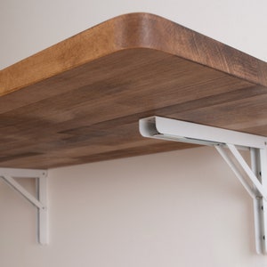 Puede incluir: Soportes plegables de metal blanco que sostienen un estante de madera. El estante est&aacute; hecho de madera marr&oacute;n claro con un patr&oacute;n de veta natural.