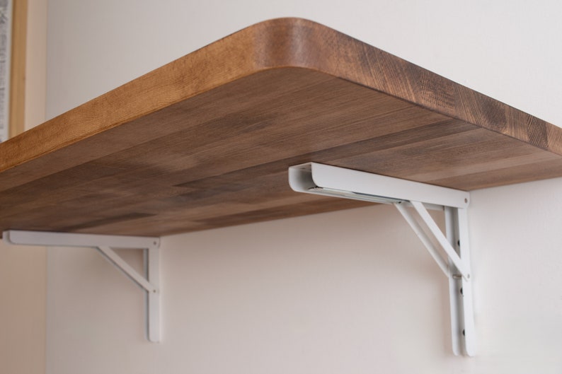 Puede incluir: Soportes plegables de metal blanco que sostienen un estante de madera. El estante est&aacute; hecho de madera marr&oacute;n claro con un patr&oacute;n de veta natural.
