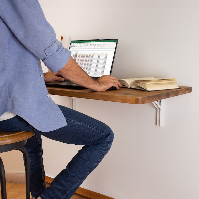 Puede incluir: Una persona se sienta en un peque&ntilde;o escritorio de pared de madera con un port&aacute;til abierto a una hoja de c&aacute;lculo. El escritorio tiene soportes de metal blanco y est&aacute; montado en una pared blanca. La persona lleva pantalones vaqueros azules y una camisa azul claro.