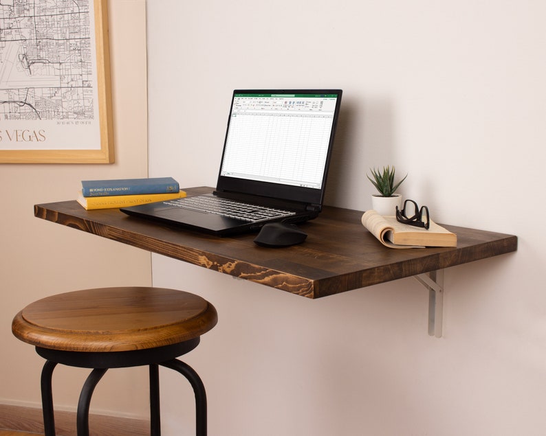 Pode incluir: Uma escrivaninha flutuante de madeira marrom escura com um laptop, livros e uma pequena planta em vaso. A escrivaninha &eacute; montada em uma parede branca com suportes de metal. Um banquinho de madeira marrom est&aacute; em primeiro plano.