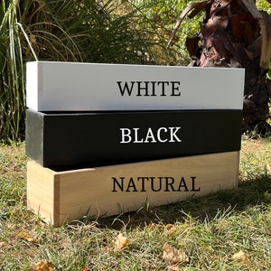 May include: Three wooden boxes stacked on top of each other. The top box is white with the word "WHITE" printed on it. The middle box is black with the word "BLACK" printed on it. The bottom box is natural wood with the word "NATURAL" printed on it.