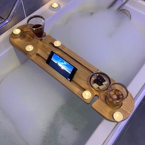May include: A wooden bathtub tray with a phone, two glasses, and several candles. The tray is resting on a bathtub filled with bubbles. One glass contains red wine, the other has ice and a brown liquid.