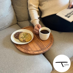 Könnte beinhalten: Ein rundes Holztablett mit leicht erhöhtem Rand. Das Tablett steht auf einem grauen Sofa und hat eine Tasse Kaffee, zwei Kekse und einen weißen Teller darauf. Das Tablett ist faltbar, wie das Schwarz-Weiß-Diagramm in der unteren rechten Ecke des Bildes zeigt.