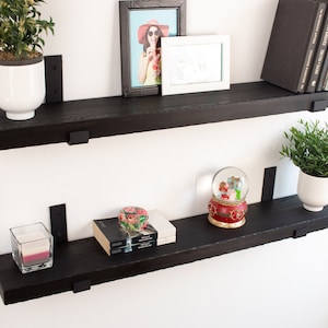 May include: Two black wooden shelves with metal brackets, displaying a variety of items, including a potted plant, a framed photo, a snow globe, books, and a candle.