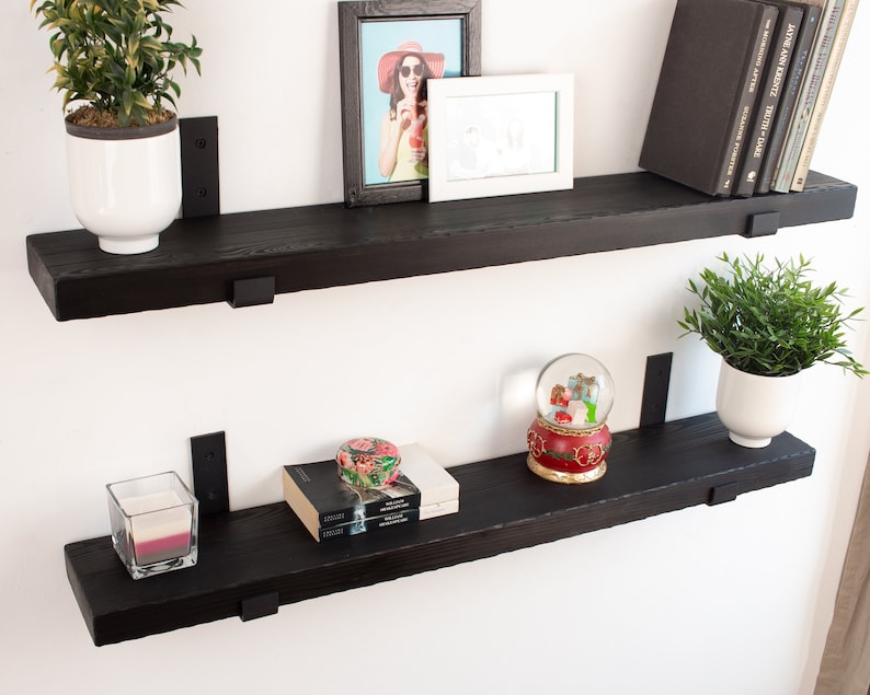 May include: Two black wooden shelves with metal brackets, displaying a variety of items, including a potted plant, a framed photo, a snow globe, books, and a candle.