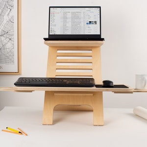 May include: A wooden standing desk with a laptop, keyboard, mouse, and a white mug on the top surface. The desk has a shelf for the keyboard and a pull-out shelf for the mouse. The desk is made of light wood and has a natural finish.