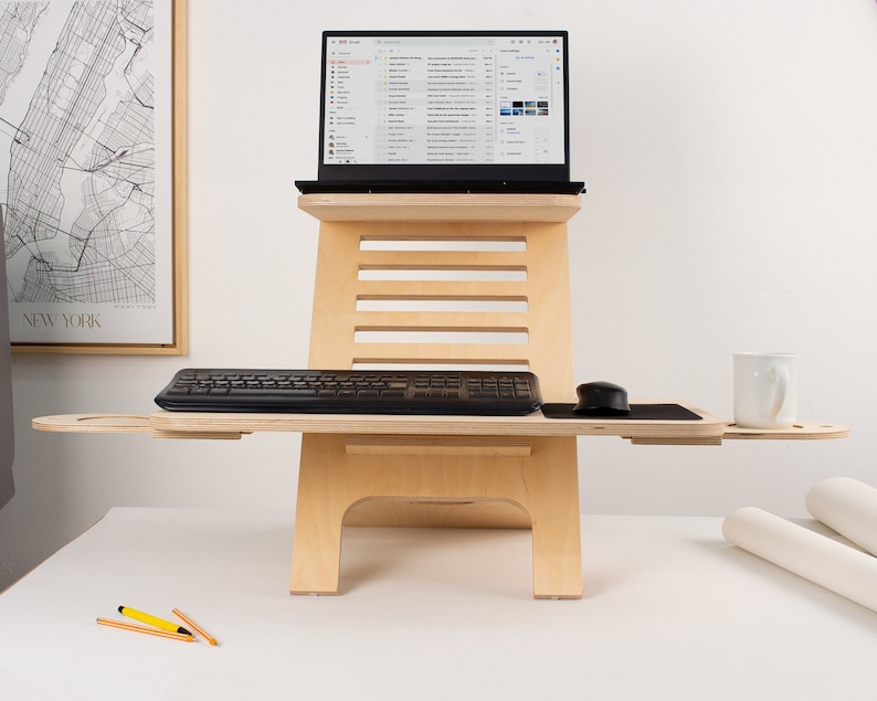 May include: A wooden standing desk with a laptop, keyboard, mouse, and a white mug on the top surface. The desk has a shelf for the keyboard and a pull-out shelf for the mouse. The desk is made of light wood and has a natural finish.