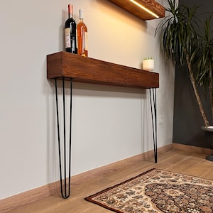 Puede incluir: Una consola de madera con acabado teñido oscuro, soportada por patas de horquilla negras. Dos botellas de vino y una pequeña planta en maceta se encuentran sobre la superficie de la mesa. La mesa está montada en una pared clara.