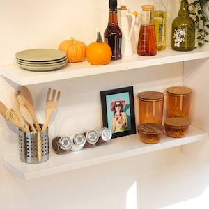 May include: Two white shelves with various kitchen items, including a set of wooden spoons, a set of glass jars with wooden lids, a framed photo of a woman wearing a hat and sunglasses, and a set of small glass jars with lids.