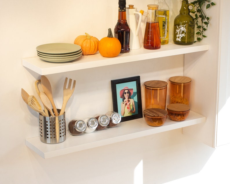 May include: Two white shelves with various kitchen items, including a set of wooden spoons, a set of glass jars with wooden lids, a framed photo of a woman wearing a hat and sunglasses, and a set of small glass jars with lids.