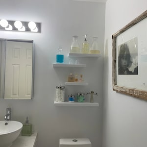 May include: A white bathroom with a white sink, a white toilet, and white shelves with various bottles and jars. There is a framed black and white photograph on the wall.