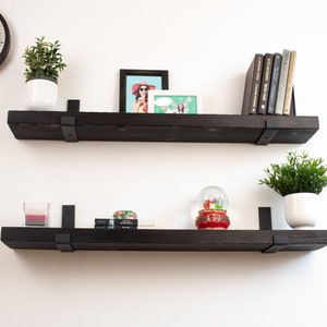 May include: Two black floating shelves with metal brackets, each shelf is decorated with plants, books, and decorative items. The top shelf has a framed photo, a potted plant, and a stack of books. The bottom shelf has a potted plant, a snow globe, and a candle.