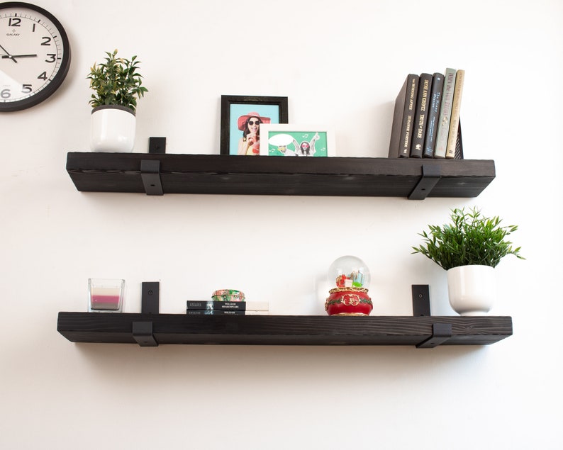May include: Two black floating shelves with metal brackets, each shelf is decorated with plants, books, and decorative items. The top shelf has a framed photo, a potted plant, and a stack of books. The bottom shelf has a potted plant, a snow globe, and a candle.