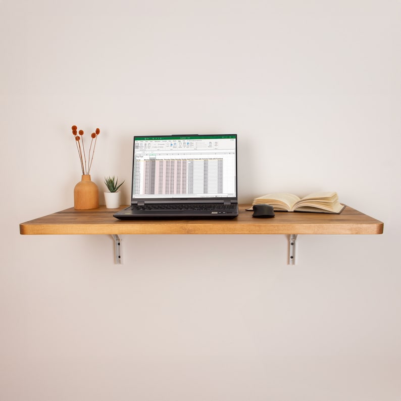 Puede incluir: Una estanter&iacute;a de madera con un ordenador port&aacute;til, un libro y un rat&oacute;n. El port&aacute;til est&aacute; abierto y muestra una hoja de c&aacute;lculo con datos. La estanter&iacute;a est&aacute; montada en una pared blanca.