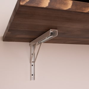 Pode incluir: Um suporte de prateleira dobr&aacute;vel de metal branco com uma prateleira de madeira marrom anexada. O suporte &eacute; fixado a uma parede com parafusos.