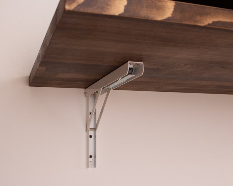 Pode incluir: Um suporte de prateleira dobr&aacute;vel de metal branco com uma prateleira de madeira marrom anexada. O suporte &eacute; fixado a uma parede com parafusos.