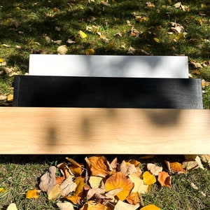 May include: Three wooden shelves, one white, one black, and one natural wood, stacked on top of each other. The shelves are sitting on a bed of fallen autumn leaves.