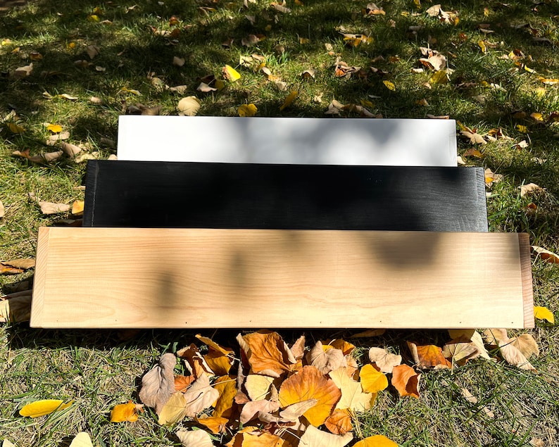 May include: Three wooden shelves, one white, one black, and one natural wood, stacked on top of each other. The shelves are sitting on a bed of fallen autumn leaves.