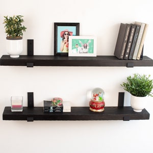 May include: Two black floating shelves with metal brackets, mounted on a white wall. The top shelf has a potted plant, a framed photo of a woman wearing a hat, and a framed photo of a couple. The bottom shelf has a potted plant, a candle, a snow globe, and a stack of books.