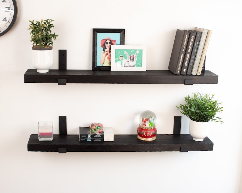 May include: Two black floating shelves with metal brackets, mounted on a white wall. The top shelf has a potted plant, a framed photo of a woman wearing a hat, and a framed photo of a couple. The bottom shelf has a potted plant, a candle, a snow globe, and a stack of books.
