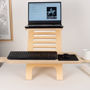 May include: A wooden standing desk with a laptop, keyboard, mouse, and a cup on a side shelf. The desk is made of light wood and has a simple design.