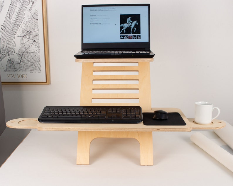 May include: A wooden standing desk with a laptop, keyboard, mouse, and a cup on a side shelf. The desk is made of light wood and has a simple design.