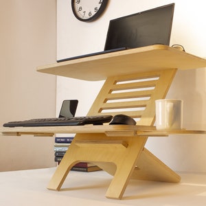May include: A light wood standing desk with a laptop, keyboard, mouse, and a white mug on the top shelf. The desk has a lower shelf for a phone and a stack of books.