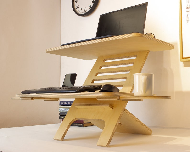 May include: A light wood standing desk with a laptop, keyboard, mouse, and a white mug on the top shelf. The desk has a lower shelf for a phone and a stack of books.