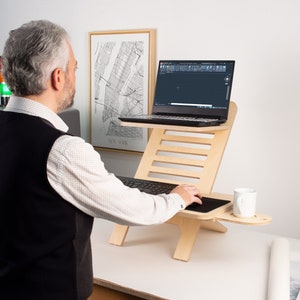 May include: A person using a laptop on a wooden standing desk. The desk has a built-in cup holder and a black mouse pad. The laptop is open and shows a black screen with a white cursor.