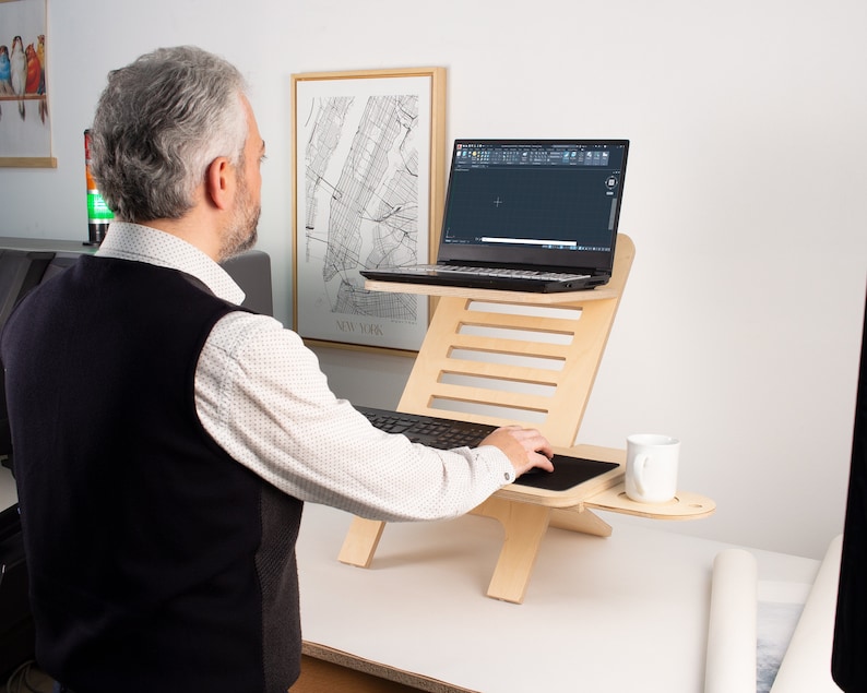 May include: A person using a laptop on a wooden standing desk. The desk has a built-in cup holder and a black mouse pad. The laptop is open and shows a black screen with a white cursor.