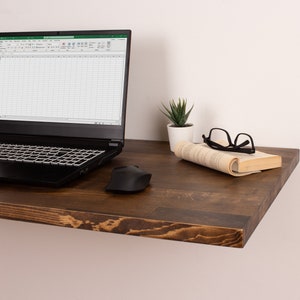Pode incluir: Uma prateleira flutuante de madeira castanho escura com um laptop, um rato sem fios, um livro, uns &oacute;culos e uma pequena planta suculenta em vaso.
