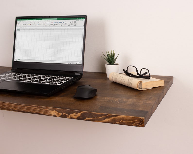 Pode incluir: Uma prateleira flutuante de madeira castanho escura com um laptop, um rato sem fios, um livro, uns &oacute;culos e uma pequena planta suculenta em vaso.