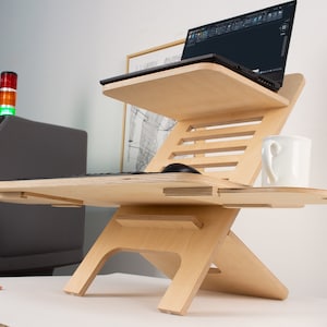 May include: A wooden standing desk with two tiers, a laptop on the top tier, a keyboard and mouse on the lower tier, and a white mug on the right side.