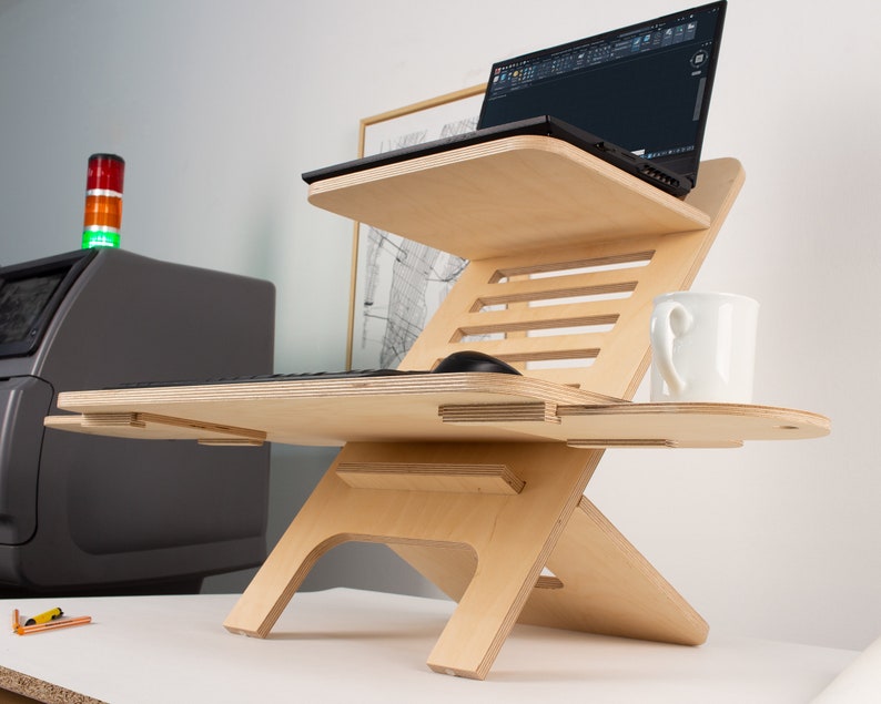 May include: A wooden standing desk with two tiers, a laptop on the top tier, a keyboard and mouse on the lower tier, and a white mug on the right side.