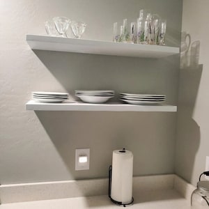 May include: Two white floating shelves with various glassware and white plates on them. The shelves are mounted on a light green wall. A white countertop with a white paper towel holder and a grey mat is below the shelves.