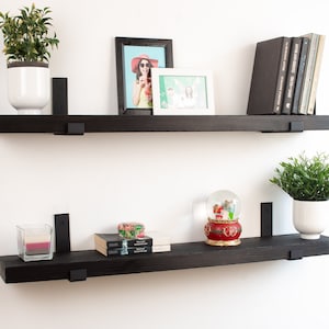 May include: Two black floating shelves with metal brackets, mounted on a white wall. The top shelf has a potted plant, a framed photo, and a stack of books. The bottom shelf has a candle, a snow globe, and a stack of books.