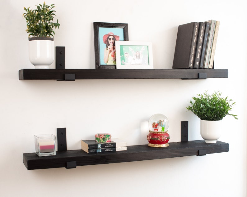 May include: Two black floating shelves with metal brackets, mounted on a white wall. The top shelf has a potted plant, a framed photo, and a stack of books. The bottom shelf has a candle, a snow globe, and a stack of books.