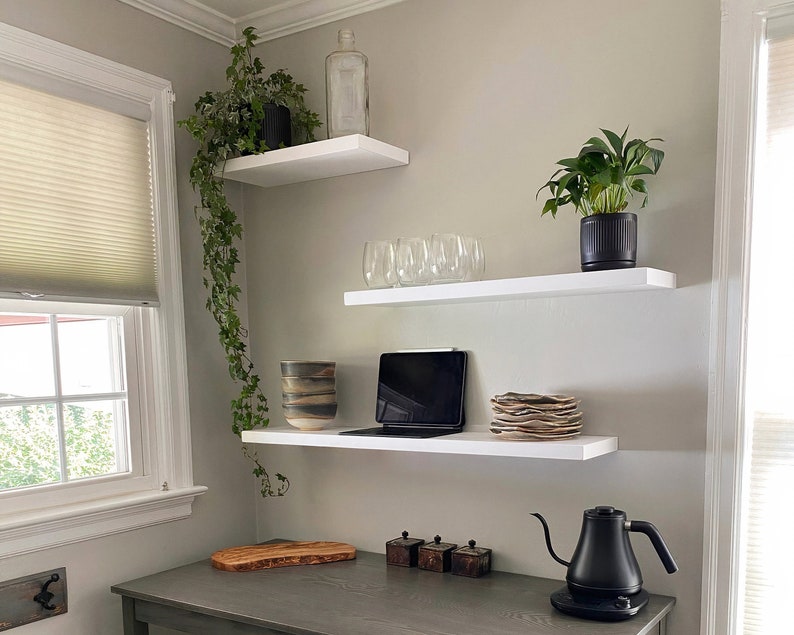 May include: Three white shelves mounted on a wall with a potted plant, a glass vase, a laptop, a stack of plates, and a black electric kettle on the bottom shelf.