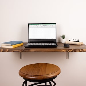 Pode incluir: Uma mesa flutuante de madeira com um laptop, uma pequena planta em vaso, um par de &oacute;culos, um livro e um rato de computador. A mesa &eacute; montada na parede e tem um banquinho de madeira por baixo.