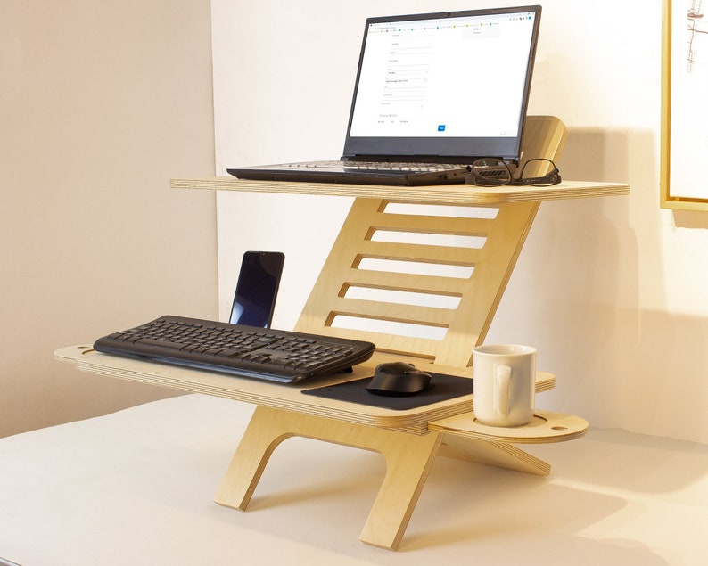 May include: A light wood standing desk with a laptop, keyboard, mouse, and a cell phone on the top level. A white mug sits on a small shelf on the bottom level.