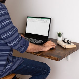 Pode incluir: Uma pessoa est&aacute; sentada em uma pequena mesa de madeira marrom escura com um laptop aberto em uma planilha. Uma pequena planta em vaso e um livro com &oacute;culos em cima est&atilde;o na mesa.