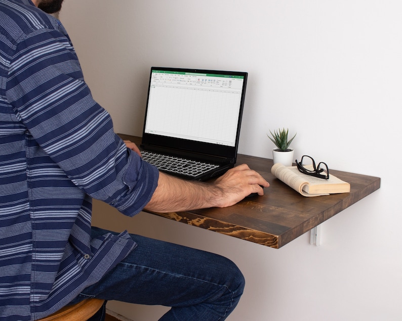 Pode incluir: Uma pessoa est&aacute; sentada em uma pequena mesa de madeira marrom escura com um laptop aberto em uma planilha. Uma pequena planta em vaso e um livro com &oacute;culos em cima est&atilde;o na mesa.
