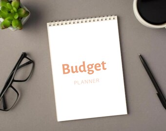 May include: A white spiral bound notebook with the text "Budget PLANNER" in orange on the front cover. The notebook is on a grey surface with a potted succulent plant, a pair of glasses, a cup of coffee, and a black pen.