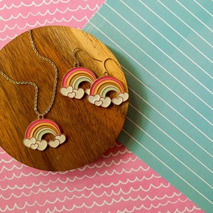 May include: A gold necklace and earrings set featuring a rainbow with white clouds charm. The rainbow is multi-coloured with pink, orange, yellow, green, blue, and purple stripes.