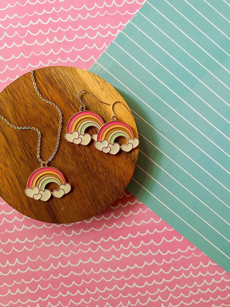 May include: A gold necklace and earrings set featuring a rainbow with white clouds charm. The rainbow is multi-coloured with pink, orange, yellow, green, blue, and purple stripes.