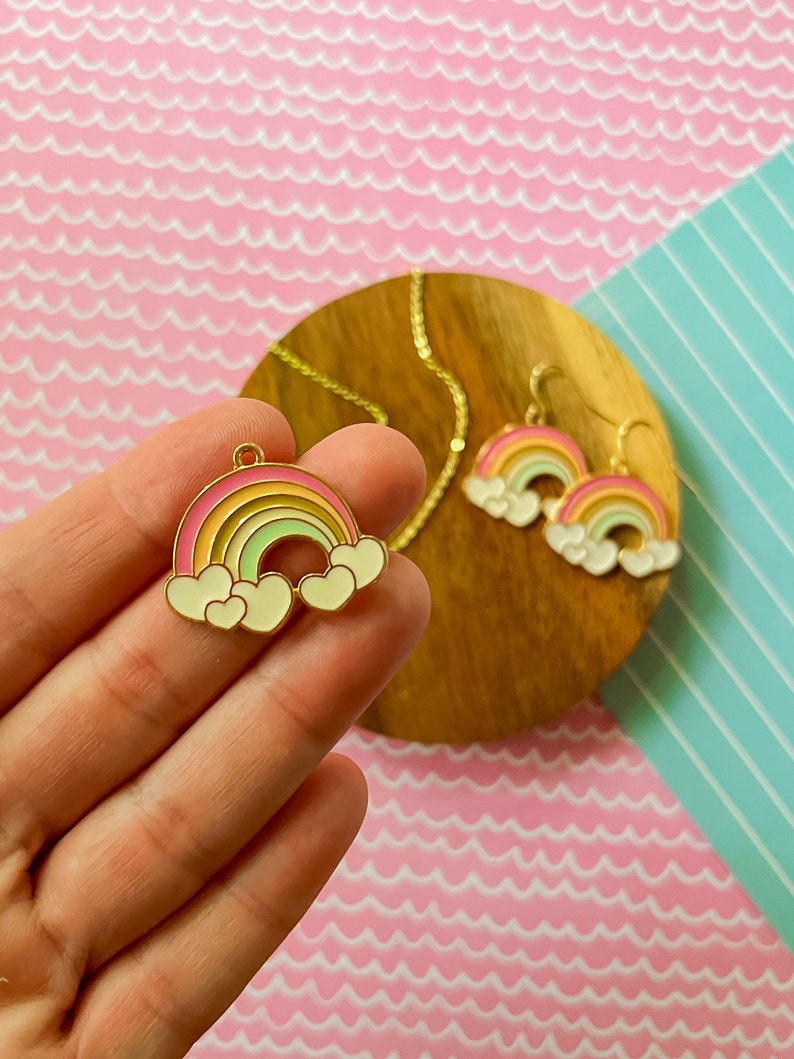 May include: A gold-tone rainbow pendant necklace and matching earrings with white clouds and hearts. The rainbow is multi-coloured with pink, yellow, and green stripes.