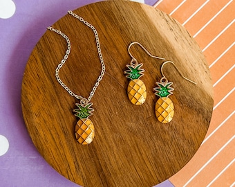May include: A gold pineapple necklace and matching earrings on a wooden surface. The pineapple charms are yellow with green tops.