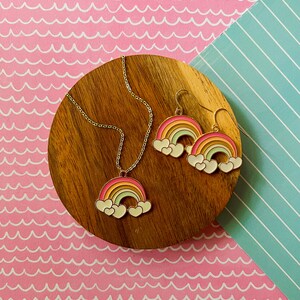May include: A gold necklace and earrings set featuring rainbow and cloud charms. The necklace has a delicate chain and the earrings are dangle style.